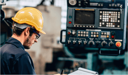 A man wearing a hard hat and glasses operates a machine in a workshop setting.